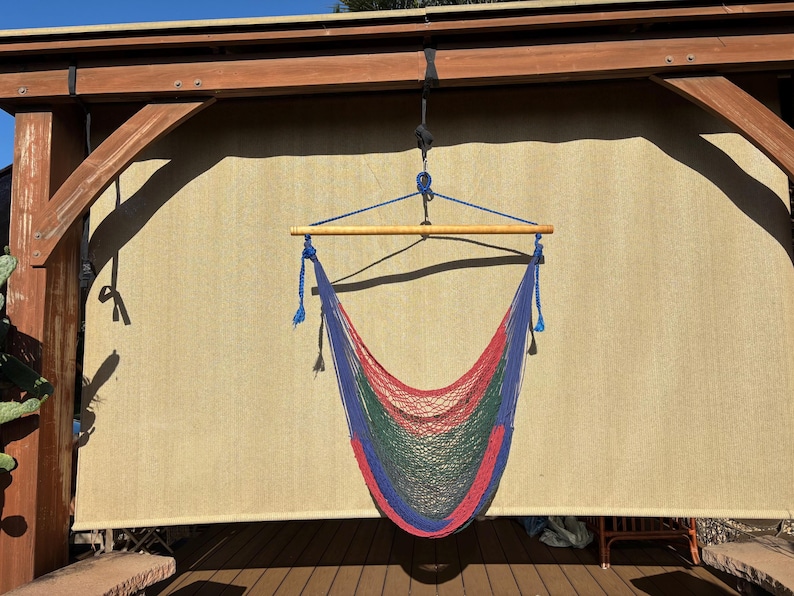 May include: A colorful hammock chair with a red, green, and blue net design, suspended from a wooden beam. The chair hangs in front of a beige backdrop, with a wooden deck below.
