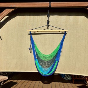 May include: A hanging hammock chair with a blue, green, and black woven net. The hammock is suspended from a wooden beam and a wooden bar. The background is a light tan shade.