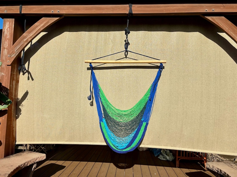 May include: A hanging hammock chair with a blue, green, and black woven net. The hammock is suspended from a wooden beam and a wooden bar. The background is a light tan shade.