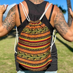 May include: A drawstring backpack with a colorful geometric pattern in shades of yellow, red, and black. The bag has white rope straps and is worn over a black tank top. The background shows a sunny outdoor scene.