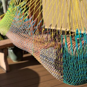 May include: A colorful hammock made of woven nylon rope. The hammock features a gradient of colors, including yellow, green, blue, and orange. The hammock is suspended, suggesting relaxation and leisure. The background includes a wooden bench and greenery.