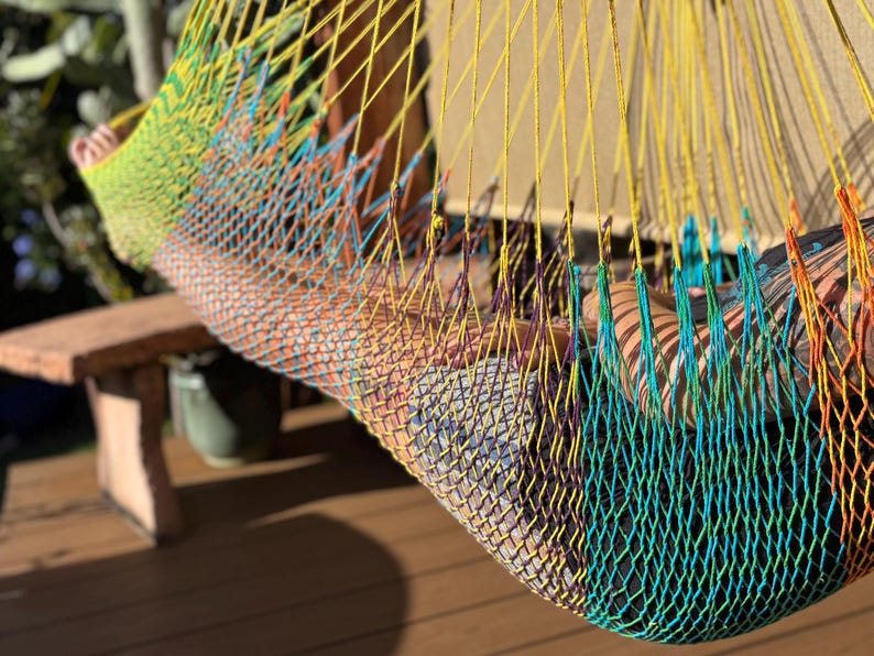 May include: A colorful hammock made of woven nylon rope. The hammock features a gradient of colors, including yellow, green, blue, and orange. The hammock is suspended, suggesting relaxation and leisure. The background includes a wooden bench and greenery.