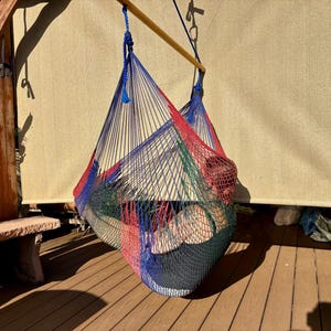 May include: A colorful hammock chair, suspended from a wooden beam, hangs on a wooden deck. The hammock is woven with red, blue, green, and black threads. A person is resting inside the hammock. The background includes a beige shade and a stone bench.