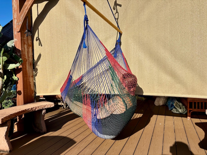 May include: A colorful hammock chair, suspended from a wooden beam, hangs on a wooden deck. The hammock is woven with red, blue, green, and black threads. A person is resting inside the hammock. The background includes a beige shade and a stone bench.