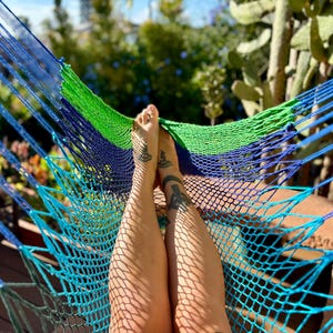 May include: A blue, green, and navy blue hammock with a woven net design. The hammock is strung between wooden supports. Legs with tattoos are resting in the hammock, with sunlight casting shadows.