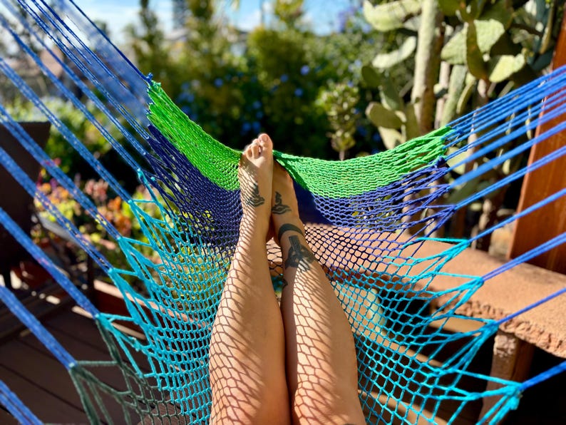 May include: A blue, green, and navy blue hammock with a woven net design. The hammock is strung between wooden supports. Legs with tattoos are resting in the hammock, with sunlight casting shadows.