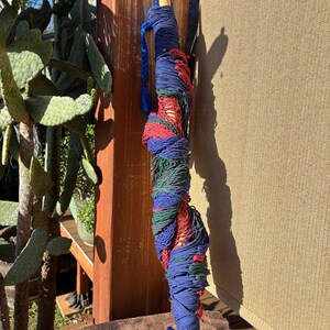 May include: A rolled-up hammock in shades of blue, red, and green, with wooden handles. The hammock is tightly wound and secured with blue rope. The wooden handles are a light brown color.