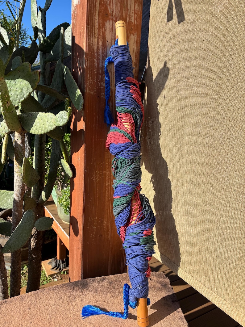 May include: A rolled-up hammock in shades of blue, red, and green, with wooden handles. The hammock is tightly wound and secured with blue rope. The wooden handles are a light brown color.