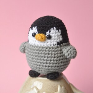 May include: A small grey and black crocheted penguin toy sitting on top of a ceramic mug with a floral design.