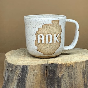 May include: White ceramic mug with a brown outline of the Adirondack Mountains and the text "ADK" in white.
