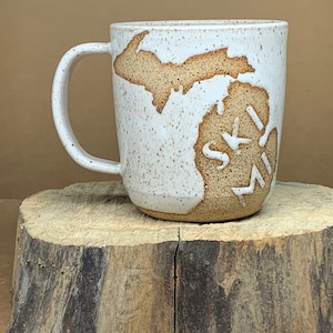 May include: White ceramic mug with a brown outline of the state of Michigan and the word "SKI MI" written inside the state outline.