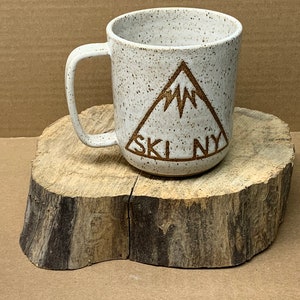 Puede incluir: Taza de cerámica blanca con un diseño de triángulo marrón y el texto "SKI NY" sobre una superficie de madera.