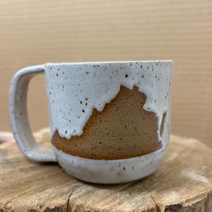 May include: A handmade ceramic mug with a white speckled glaze and a brown band featuring a state outline. The mug has a curved handle and sits on a wooden surface. The mug is a unique piece of drinkware.