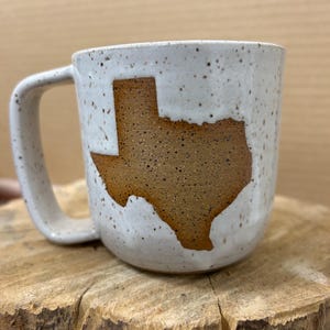 May include: A speckled white ceramic mug with a brown Texas outline. The mug has a large handle and is sitting on a wooden surface. The mug is a unique and decorative item.