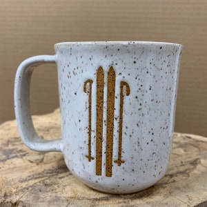 May include: A speckled white ceramic mug with a brown handle. The mug features a brown graphic of skis and ski poles. The mug is sitting on a wooden surface.
