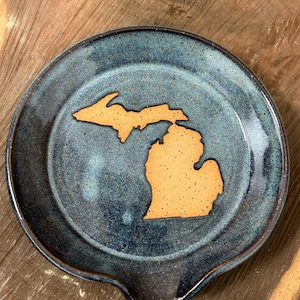 May include: A handmade ceramic spoon rest with a blue-green glaze and a tan-colored Michigan state outline in the center. The spoon rest has a curved indentation for resting utensils.
