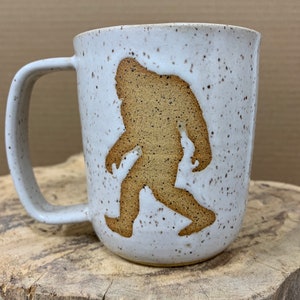 May include: White ceramic mug with a brown silhouette of Bigfoot walking on the side. The mug has a speckled glaze.