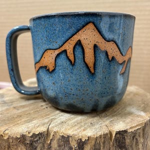 May include: A ceramic mug with a blue speckled glaze and a brown mountain range design. The mug has a dark blue handle and is sitting on a wooden surface. The mug is approximately 4 inches tall.