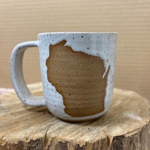 May include: A speckled white ceramic mug with a brown Wisconsin state outline. The mug has a curved handle and sits on a wooden surface. The mug is a handmade item.