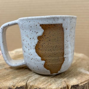 May include: A speckled white ceramic mug with a brown Illinois state outline. The mug has a curved handle and sits on a wooden surface. The interior of the mug is not visible.