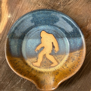 May include: A handmade ceramic spoon rest with a Bigfoot silhouette. The spoon rest features a blue and brown glaze with a Bigfoot figure in the center. The spoon rest has a small lip for holding utensils and is a unique kitchen accessory.