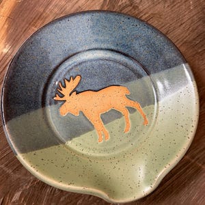 May include: A ceramic spoon rest with a moose silhouette. The spoon rest is round with a blue and green speckled glaze. The moose is a light orange color. The rest has a curved indentation for holding a spoon.