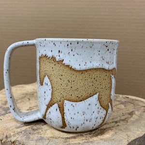 May include: A speckled white ceramic mug with a brown horse silhouette. The mug has a curved handle and a wide, cylindrical shape. The horse design is centered on the mug's body, with the background a contrasting brown color. The mug is sitting on a wooden surface.