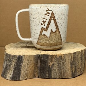 May include: White ceramic mug with a brown mountain design and the text "SKI NY" on the side. The mug is sitting on a wooden surface.