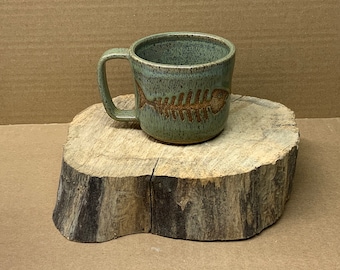 May include: A handmade ceramic mug with a seafoam green glaze and a brown fish skeleton design. The mug has a sturdy handle and is approximately 4 inches tall. The mug is sitting on a wooden slice.