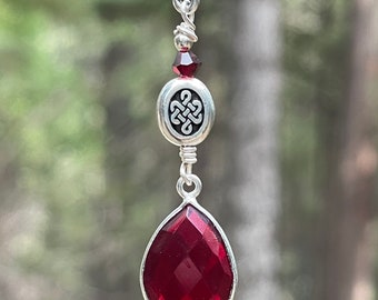May include: A silver chain necklace with a red gemstone pendant. The pendant features a silver Celtic knot design and a small silver bead.
