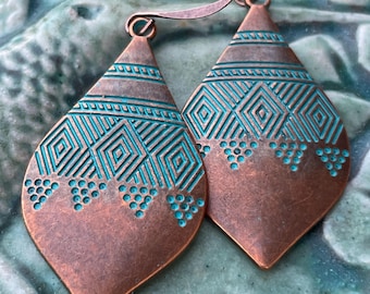 May include: A pair of copper-colored teardrop earrings with turquoise geometric patterns. The earrings have a detailed design with etched lines and small dot accents, and are suspended from curved ear wires. The earrings are displayed on a textured, teal-colored surface.