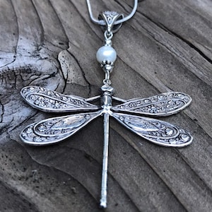 May include: A silver dragonfly pendant necklace with a white pearl accent. The dragonfly is detailed with intricate floral designs.