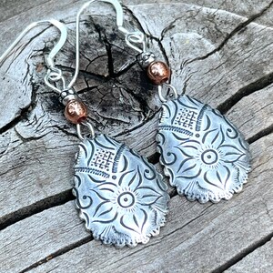 May include: A pair of silver earrings with a floral design and copper accents. The earrings are teardrop shaped and have a delicate, intricate design.