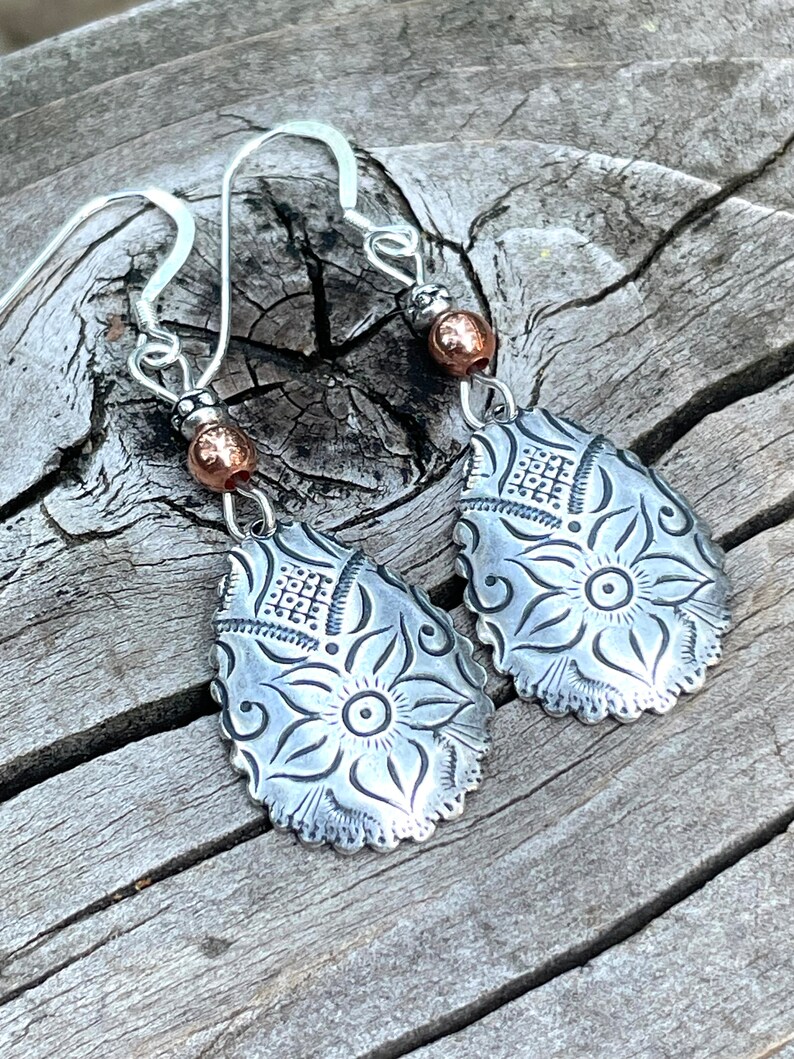 May include: A pair of silver earrings with a floral design and copper accents. The earrings are teardrop shaped and have a delicate, intricate design.