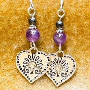 Pode incluir: Um par de brincos de prata em forma de cora&ccedil;&atilde;o com um design floral. Cada brinco tem uma conta de ametista roxa e uma conta preta.
