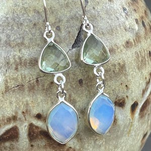 May include: A pair of dangle earrings with a silver finish. Each earring features a light green, triangular gemstone above a light blue, oval-shaped gemstone. The earrings are suspended from a curved silver hook.