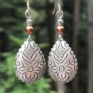 May include: A pair of silver dangle earrings with a copper bead and a floral design. The earrings are against a dark green background.
