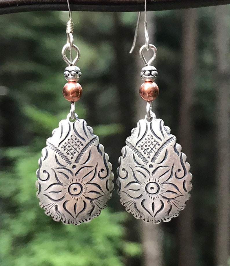 May include: A pair of silver dangle earrings with a copper bead and a floral design. The earrings are against a dark green background.