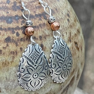 May include: Silver dangle earrings with a floral design and copper beads. The earrings are teardrop shaped and are attached to silver hooks.