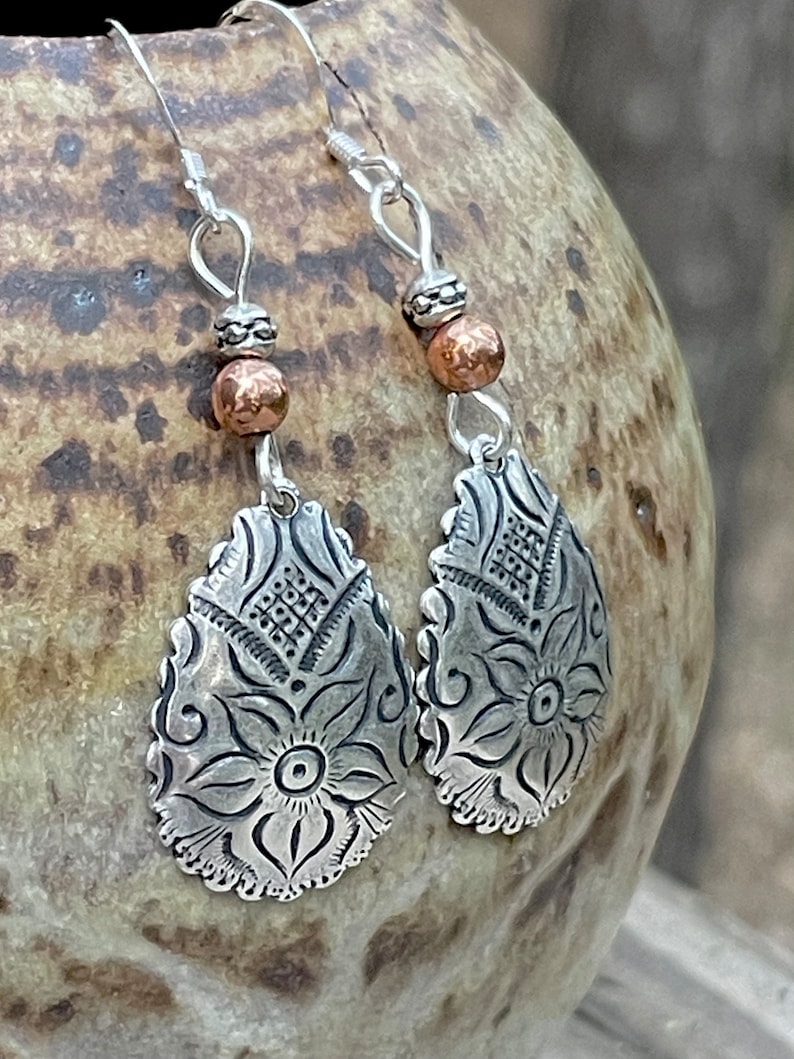 May include: Silver dangle earrings with a floral design and copper beads. The earrings are teardrop shaped and are attached to silver hooks.