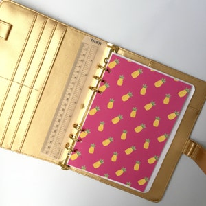 Puede incluir: Una agenda dorada abierta, con una regla transparente y una página rosa con un patrón de piñas amarillas. El interior revela anillos dorados y bolsillos. La palabra "TODAY" es visible en la regla. Un artículo de papelería.