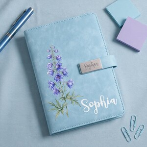May include: A light blue notebook featuring a floral illustration of purple delphinium flowers and the name Sophia printed in white script. The notebook includes a silver metal closure tab engraved with the name Sophia in cursive script.