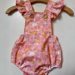 May include: Pink baby romper with a bunny rabbit print. The romper has a square neckline, ruffled sleeves, and wooden buttons. The fabric has a grid pattern and is hanging on a wooden hanger.