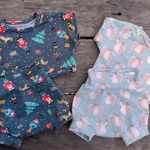 May include: Two sets of children's clothing, one with a dark blue background and a pattern of nutcrackers, Christmas trees, and gifts. The other set has a light blue background with a pattern of angels. Both sets include a short-sleeved top and shorts.