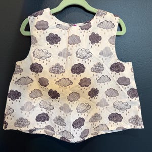 May include: A sleeveless top with a white background and a pattern of dark grey clouds with rain drops. The clouds have various textures and shapes. The top is hanging on a green hanger.