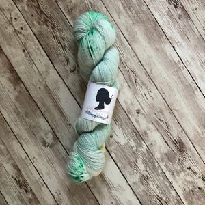 Può includere: Un gomitolo di filato tinto a mano nei toni del verde menta e del bianco, con vivaci macchie verdi. Il filato è etichettato con un'etichetta bianca con una silhouette e un motivo floreale. Il filato è esposto su una superficie di legno chiaro.