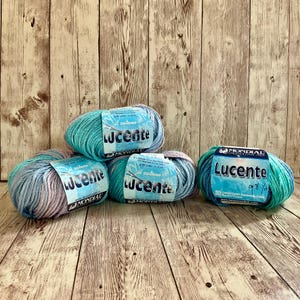 May include: Five skeins of Lucente yarn in shades of blue, teal, and lavender. Each skein has a label with the brand name Mondial and the word "Lucente". The yarn is displayed on a wooden background.
