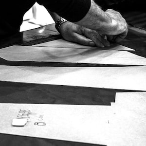 May include: Black and white photo of a person cutting out fabric pieces for a sewing project. The fabric pieces are laid out on a dark surface. The person is wearing a black shirt and has their hands on the fabric.