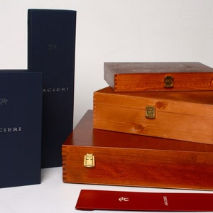May include: A set of four wooden boxes with brass clasps, stacked on top of each other. The boxes are all the same size and shape, and they are made of a light brown wood. The boxes are labeled with the word "ARCIERI" in silver lettering. A red envelope with the same logo is in the foreground.
