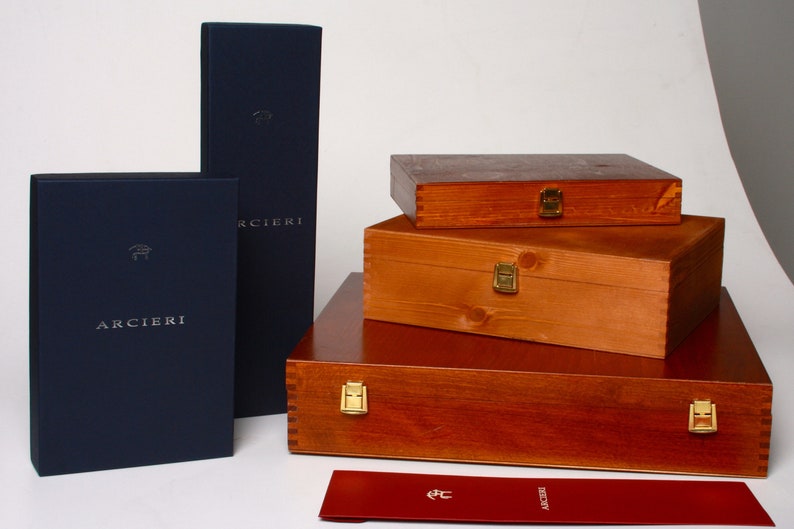 May include: A set of four wooden boxes with brass clasps, stacked on top of each other. The boxes are all the same size and shape, and they are made of a light brown wood. The boxes are labeled with the word "ARCIERI" in silver lettering. A red envelope with the same logo is in the foreground.